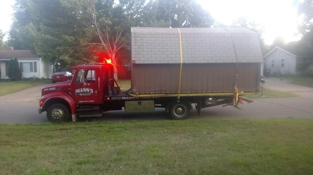 Red flatbed truck hauling a large shed or cabin on a grassy roadside, with a red warning light flashing.