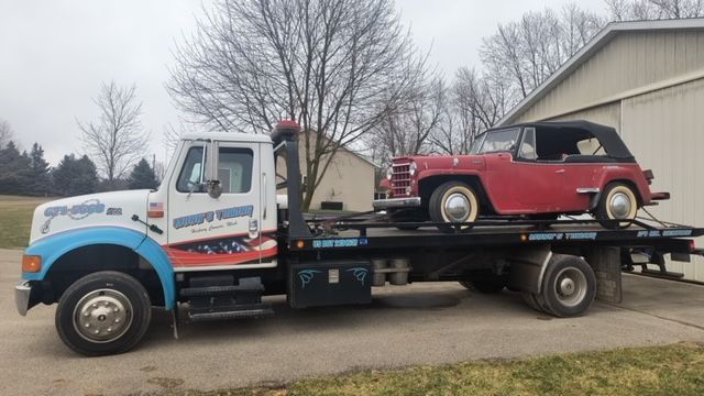 Tow truck carrying a red vintage car beside a building on an overcast day