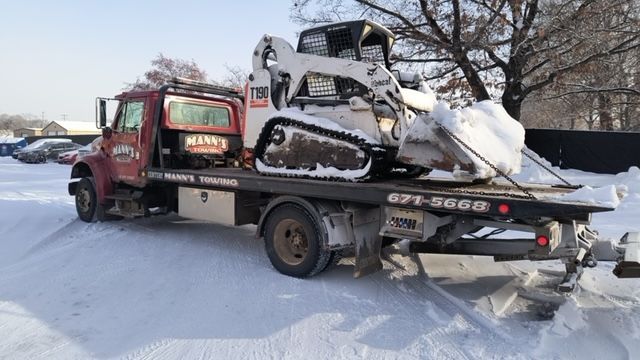 Red tow truck carrying a snow-covered skid steer on a snowy road