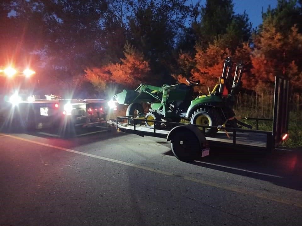 Tractor on a trailer being towed at night, with flashing emergency lights on a roadside.