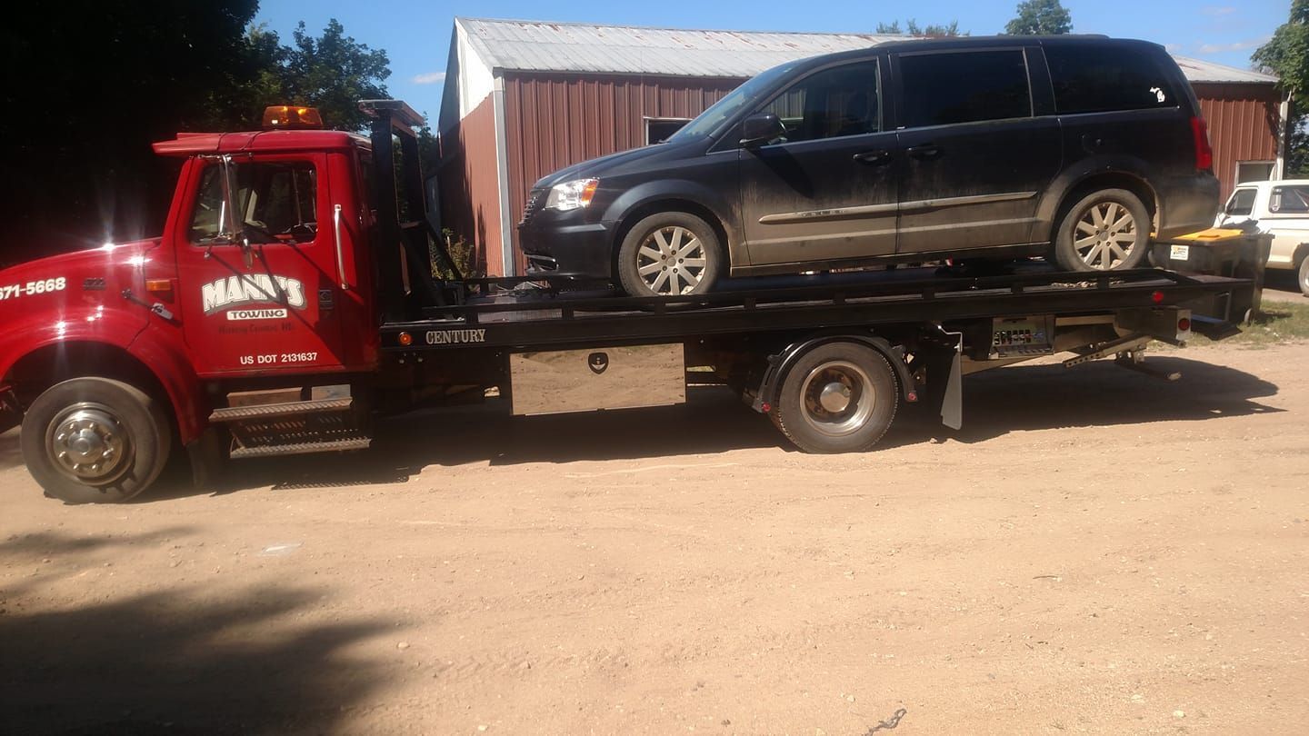 Red tow truck carrying a black SUV on a flatbed in a dirt lot near a metal building.