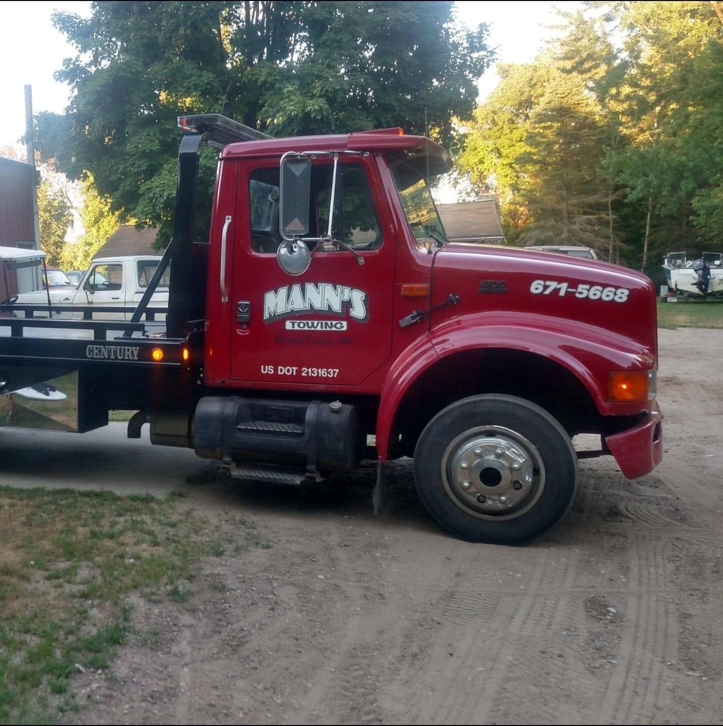 Red tow truck parked on a dirt road beside trees, with “MANN’S” on the door.