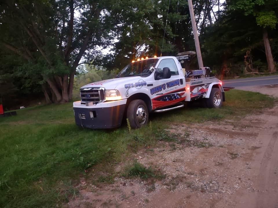 Tow truck parked on a grassy roadside at dusk, with trees and a curving road behind it.