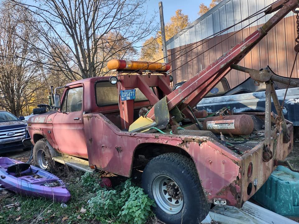 Old red tow truck with crane arm parked beside a barn, surrounded by overgrown plants and scrap debris.