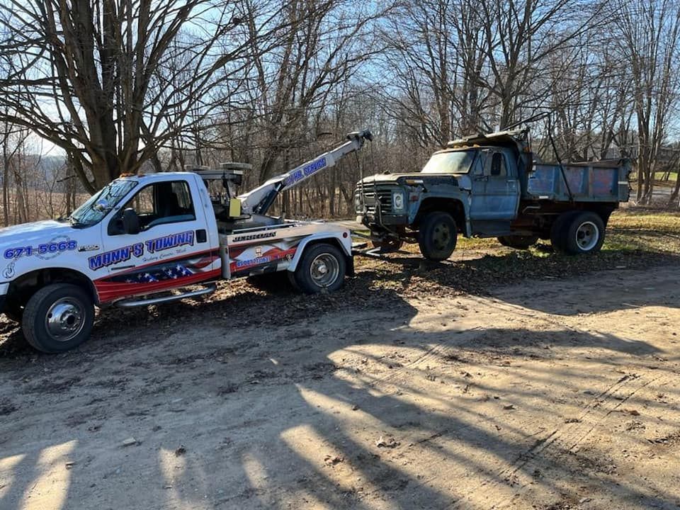 Two tow trucks parked on a dirt lot beside leafless trees, one white and one dark green.
