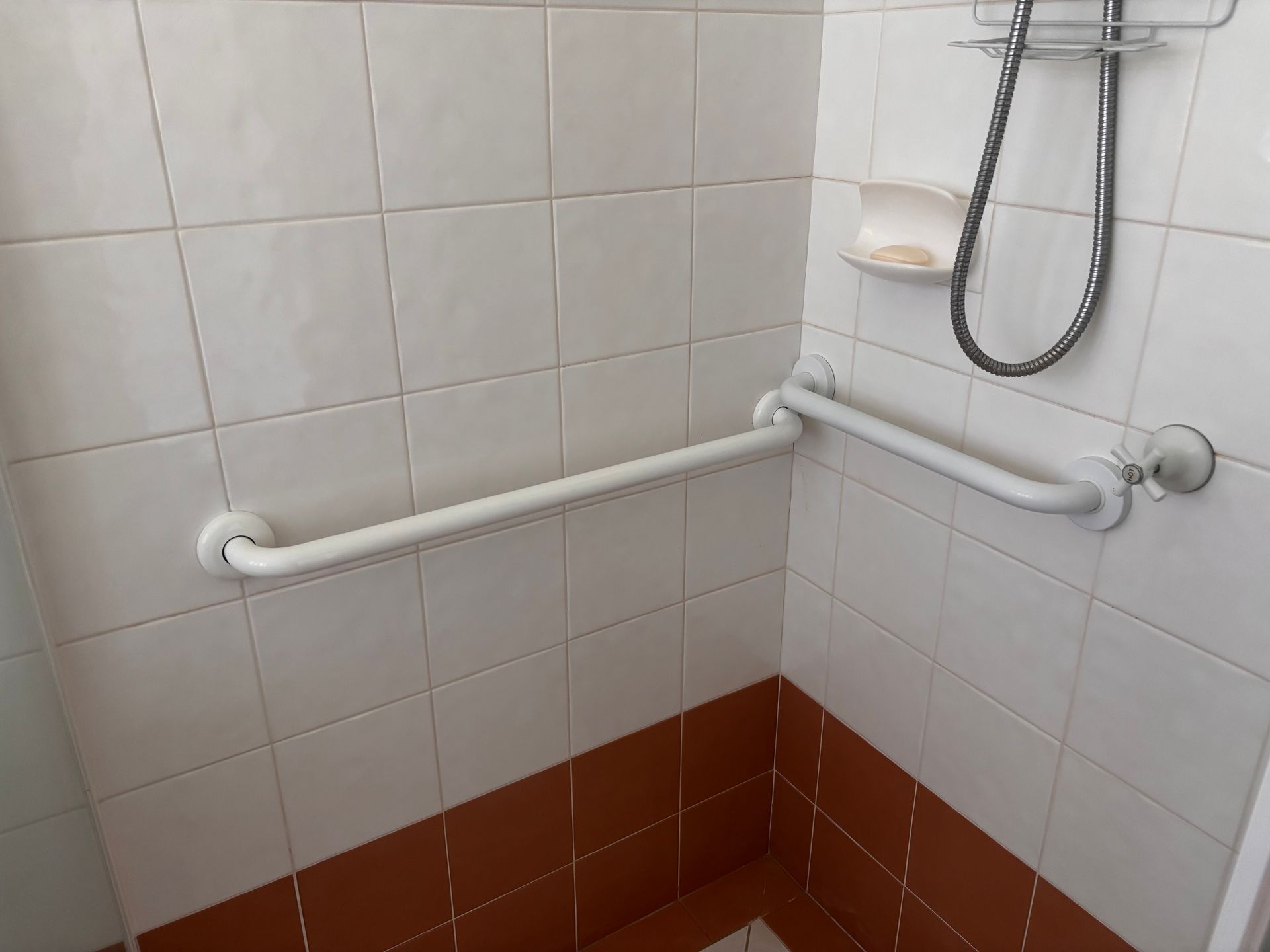 White L-shaped grab bar mounted on a white and brown tiled shower wall below a showerhead and soap dish.