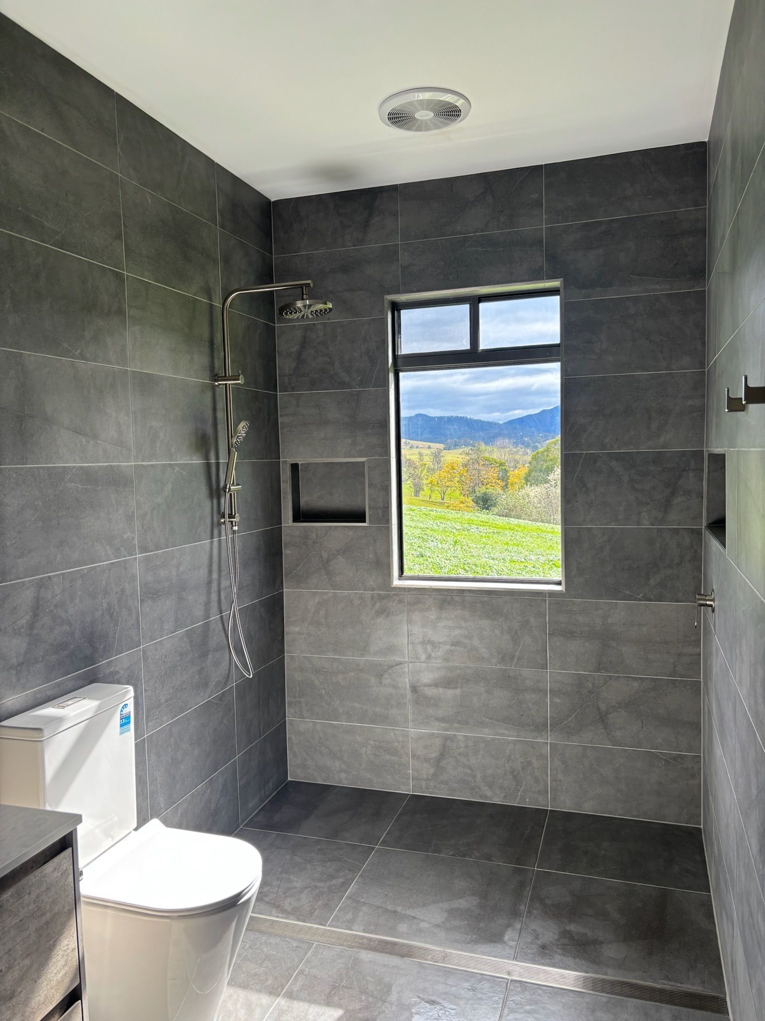 Gray Tiled Bathroom With Shower Fixture, Toilet, Window With a View — Edmonds Bathroom Renovations & Tiling In Wauchope, NSW