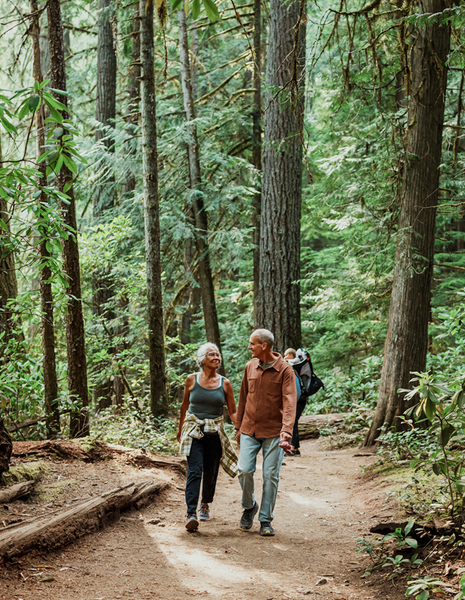 Older couple walking on a wooded path, holding hands. Trees surround them.
