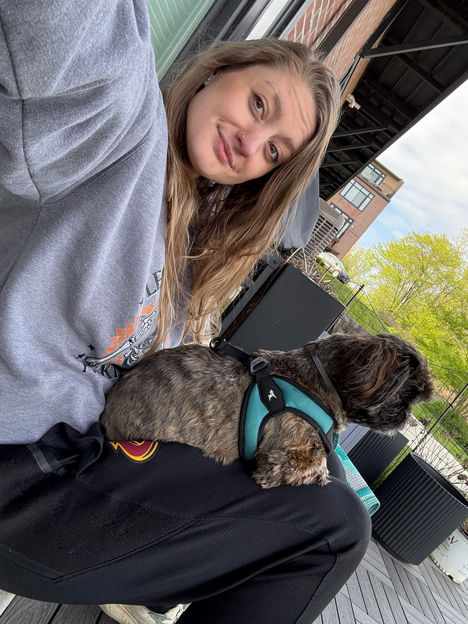 Lex smiling, sitting with small dog on lap. Outdoors, gray sweatshirt, black pants, teal harness.