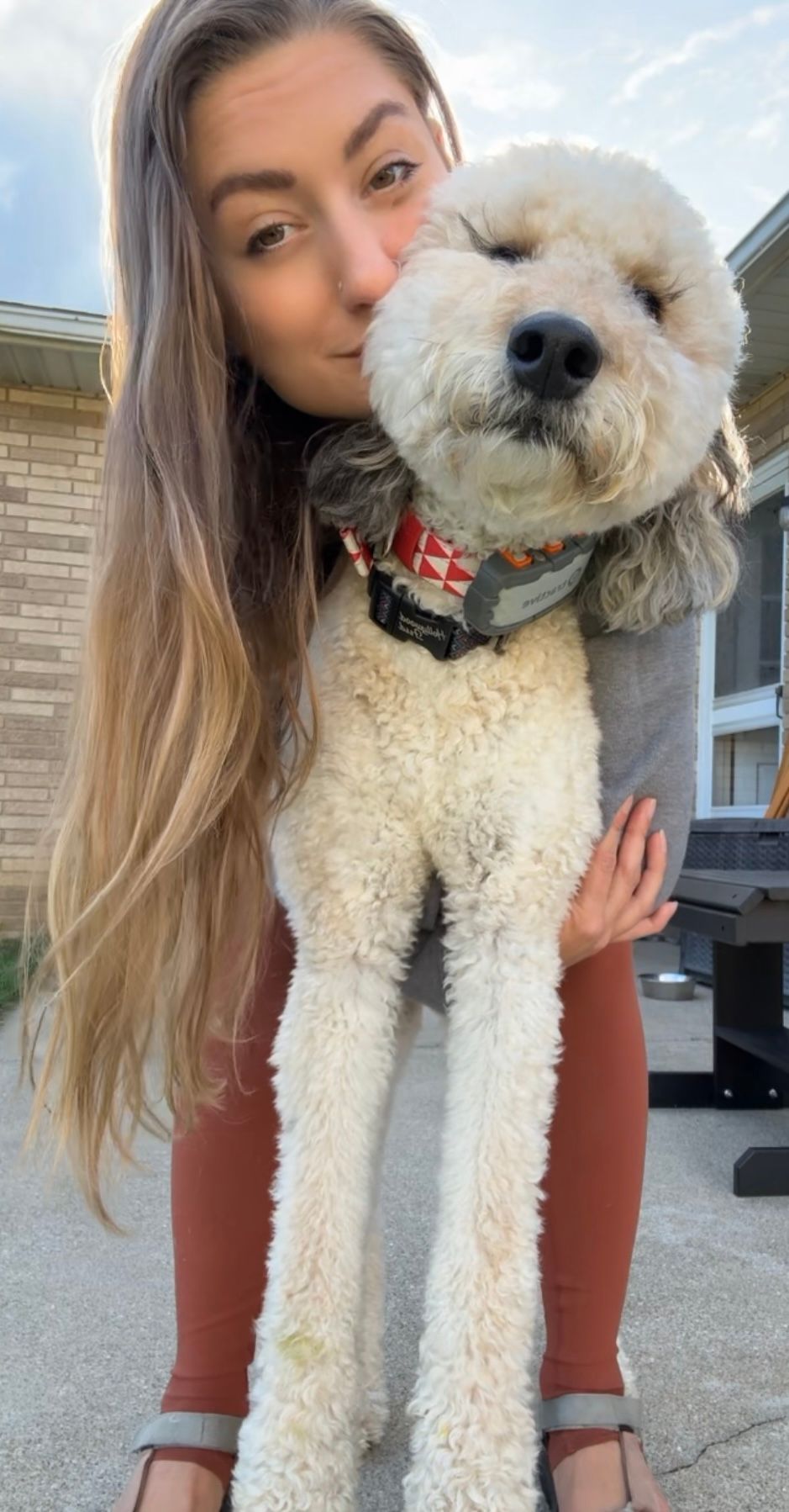 Lex holding a fluffy white dog outdoors, kissing its face. The dog has a harness and a red collar.