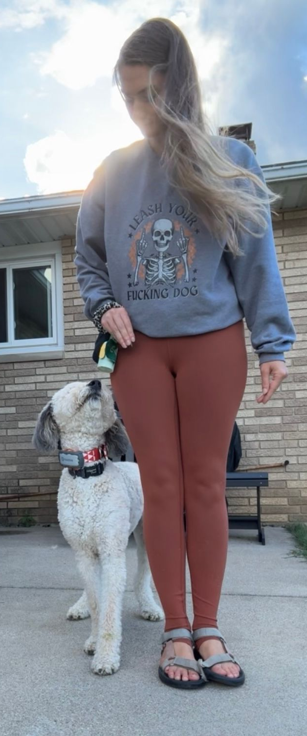 Lex and fluffy dog outside, Lex is looking down, wearing gray sweatshirt, brown leggings, sandals.