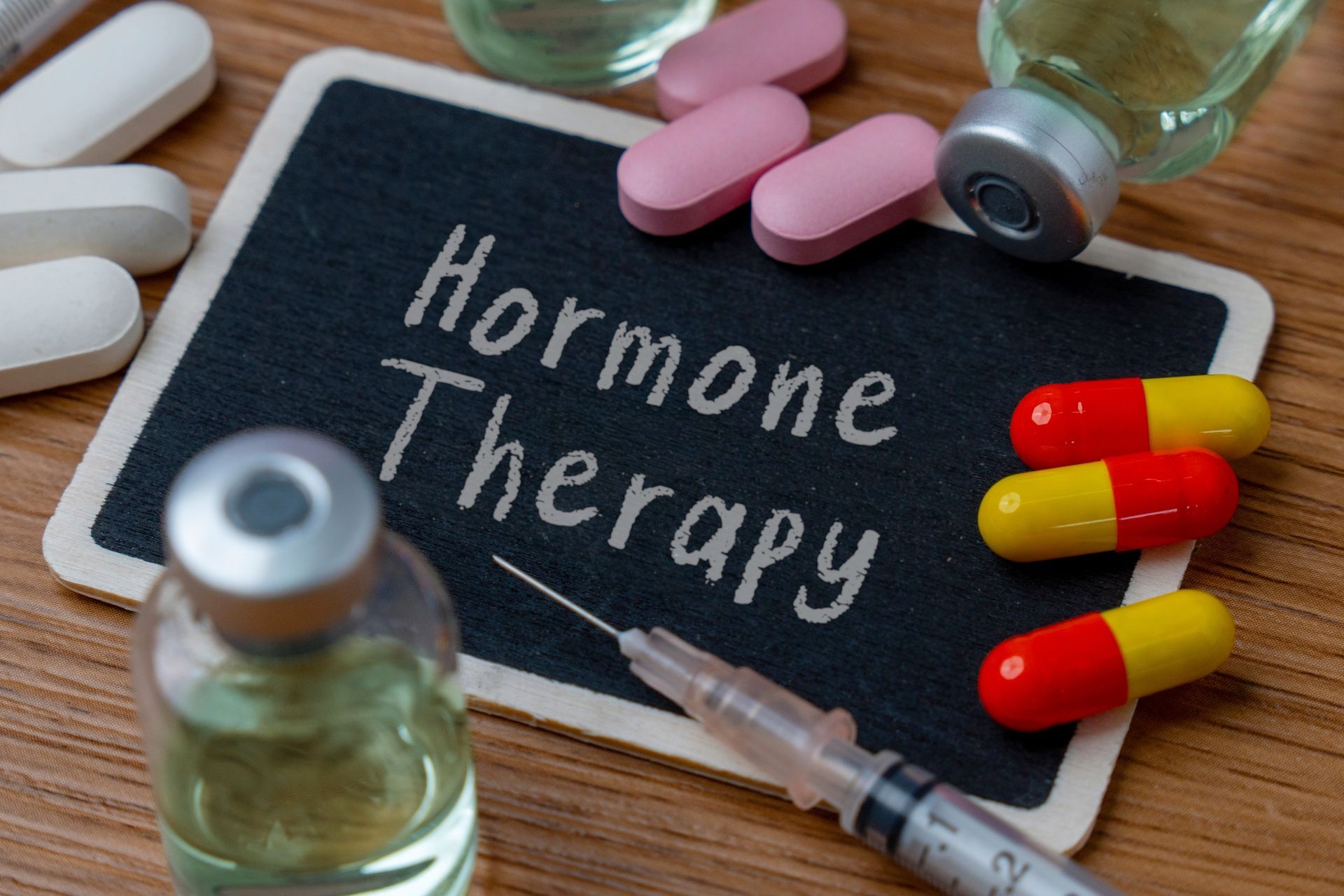 Small chalkboard with the words “Hormone Therapy,” with pills, capsules, vials, & a syringe.