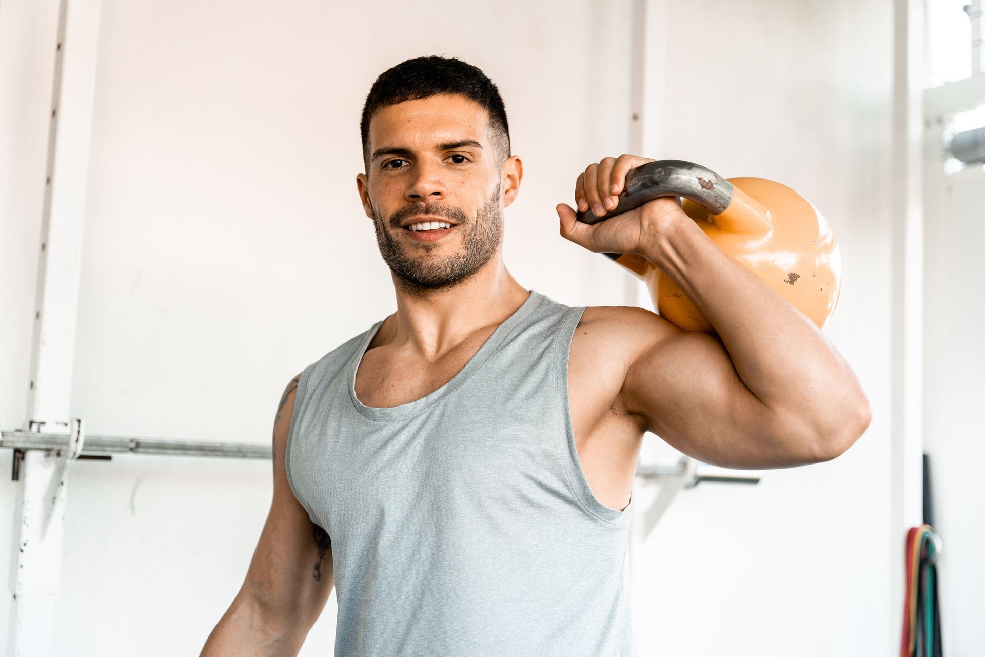Man smiling, holding a yellow kettlebell on his shoulder at a gym.