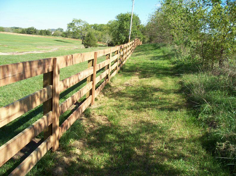 A wooden four-rail fence runs along a lush green pasture under a clear blue sky.