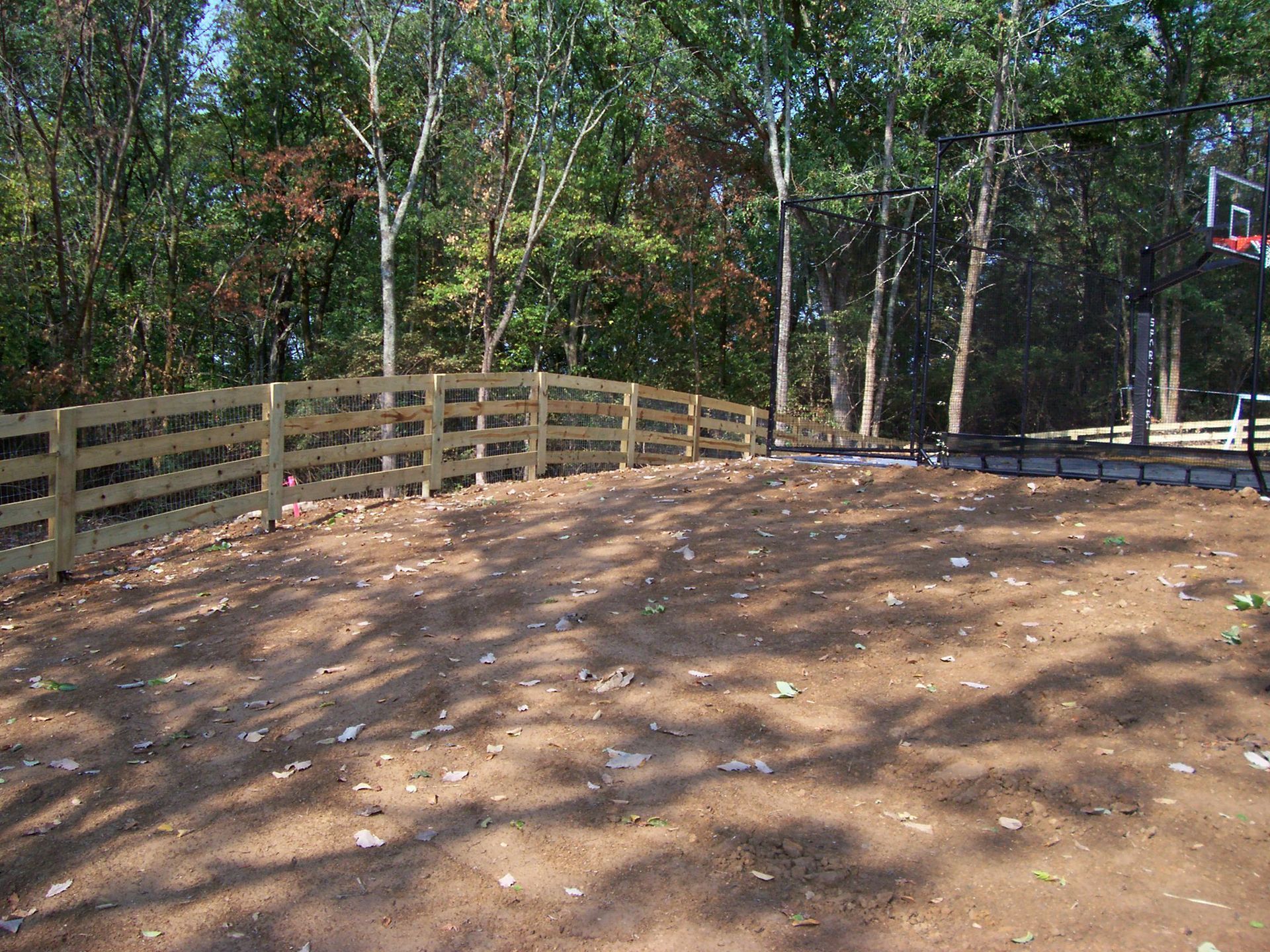A wooden split-rail fence runs along a curving, dirt backyard bordering a wooded area with a basketball hoop.