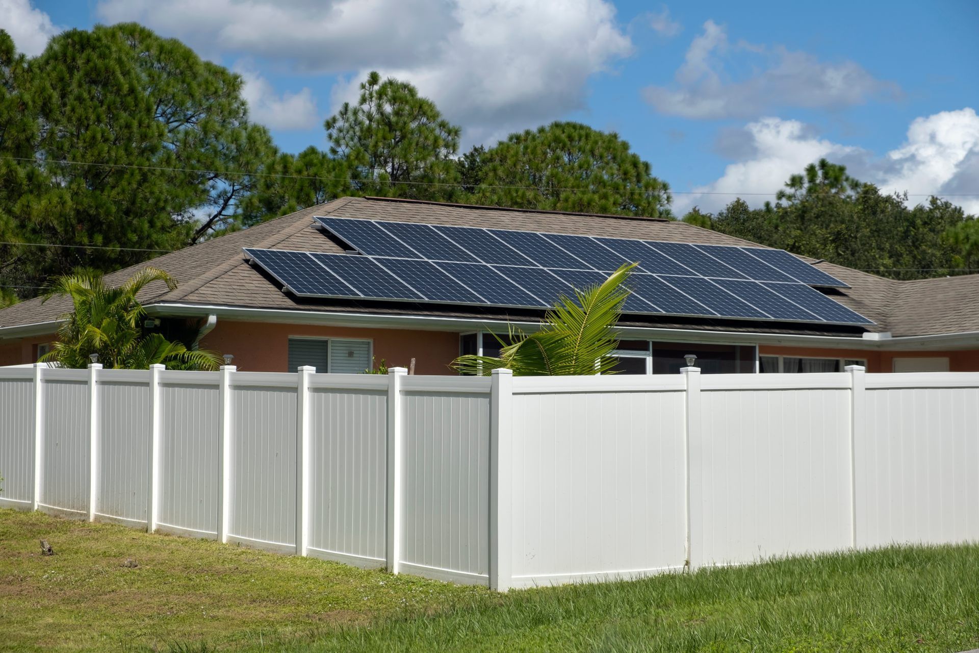 A one-story home with a tan exterior and a dark shingled roof featuring solar panels, enclosed by a white vinyl fence.