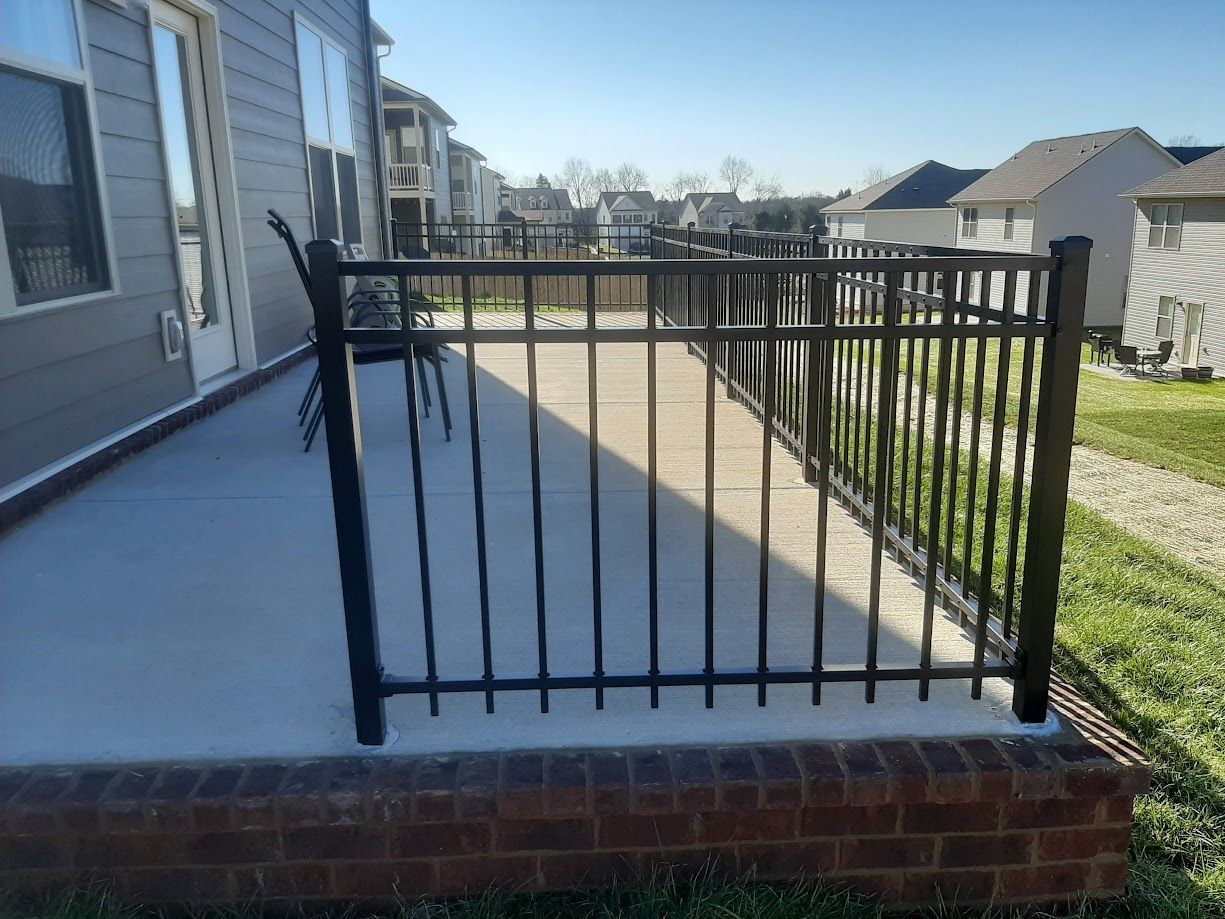 A concrete patio with black metal railings, a brick border, and chairs, located behind a suburban house on a sunny day.