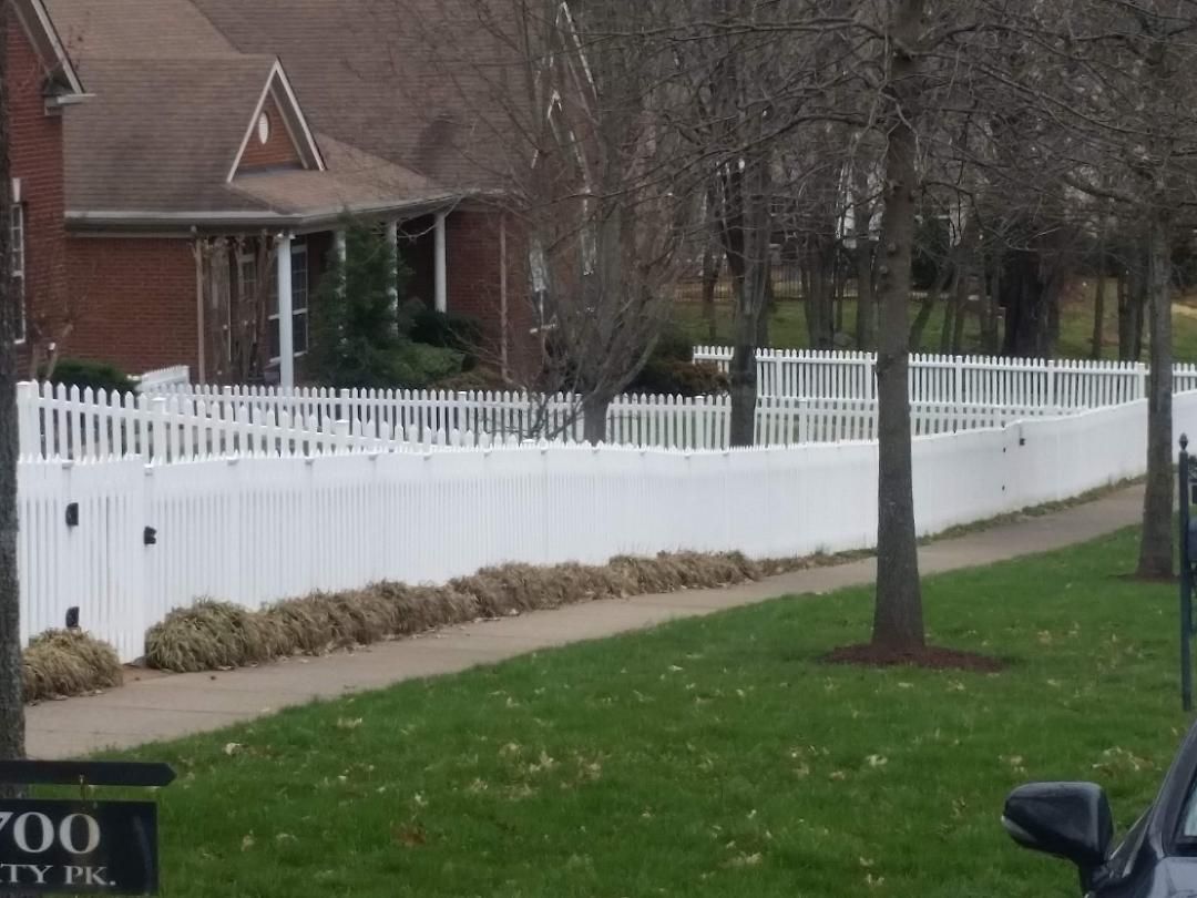 A white picket fence runs along a sidewalk in front of a brick house with bare trees in the yard.