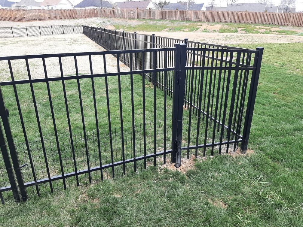 A black metal fence encloses a green grassy area with a sandy patch in the background under a cloudy sky.