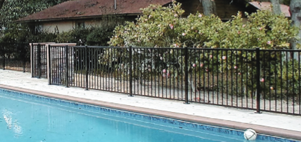 A black metal safety fence runs along the edge of a backyard swimming pool next to shrubs and a house.