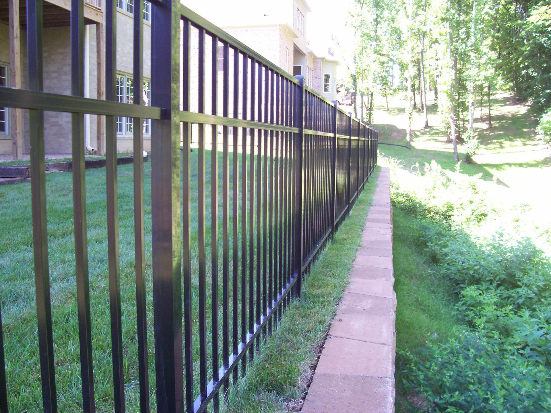 Black metal fence bordering a manicured lawn and a stone retaining wall leading toward a residential home.