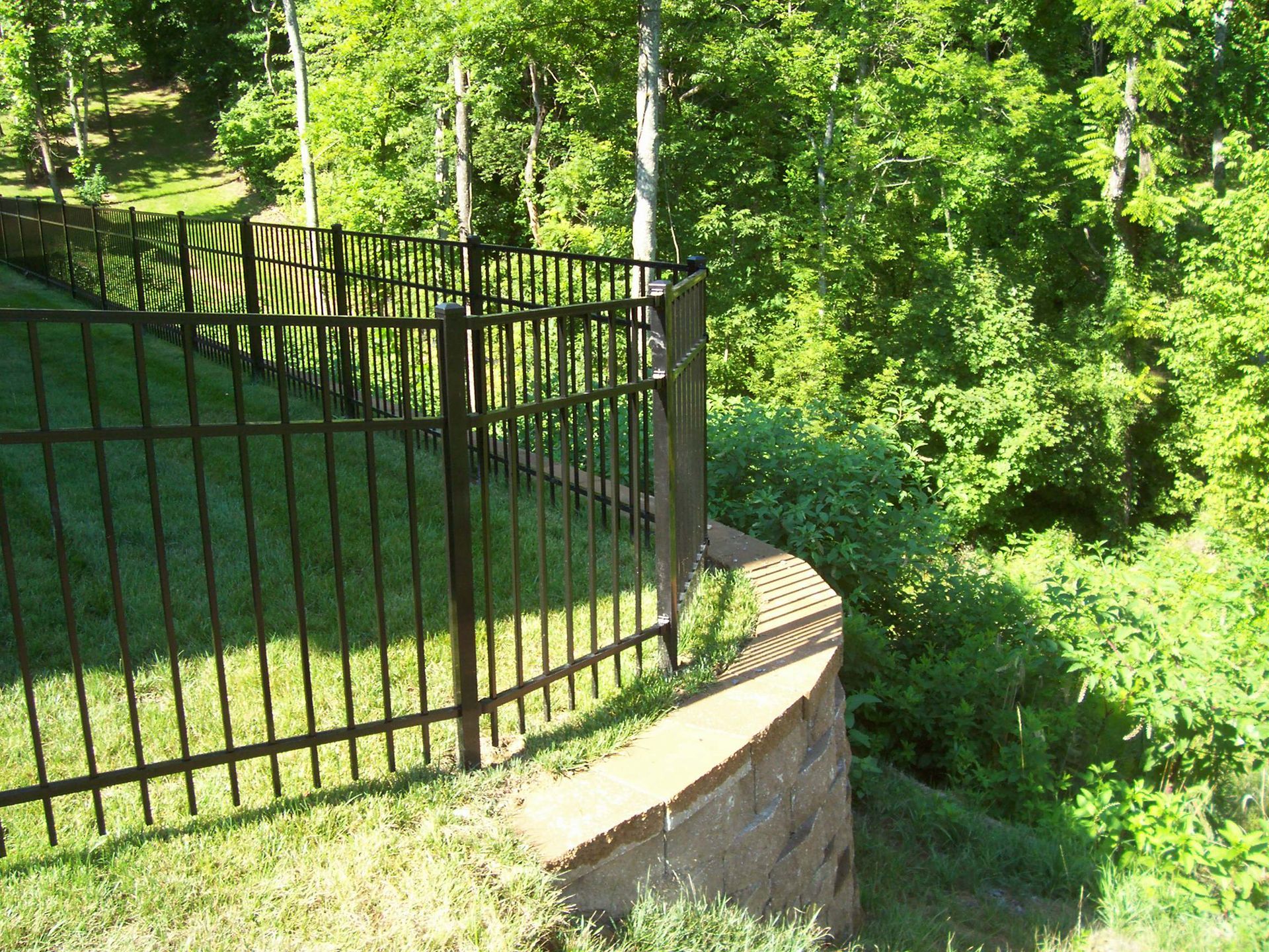 A dark metal fence lines the top of a stone retaining wall, bordering a grassy yard overlooking a wooded area.