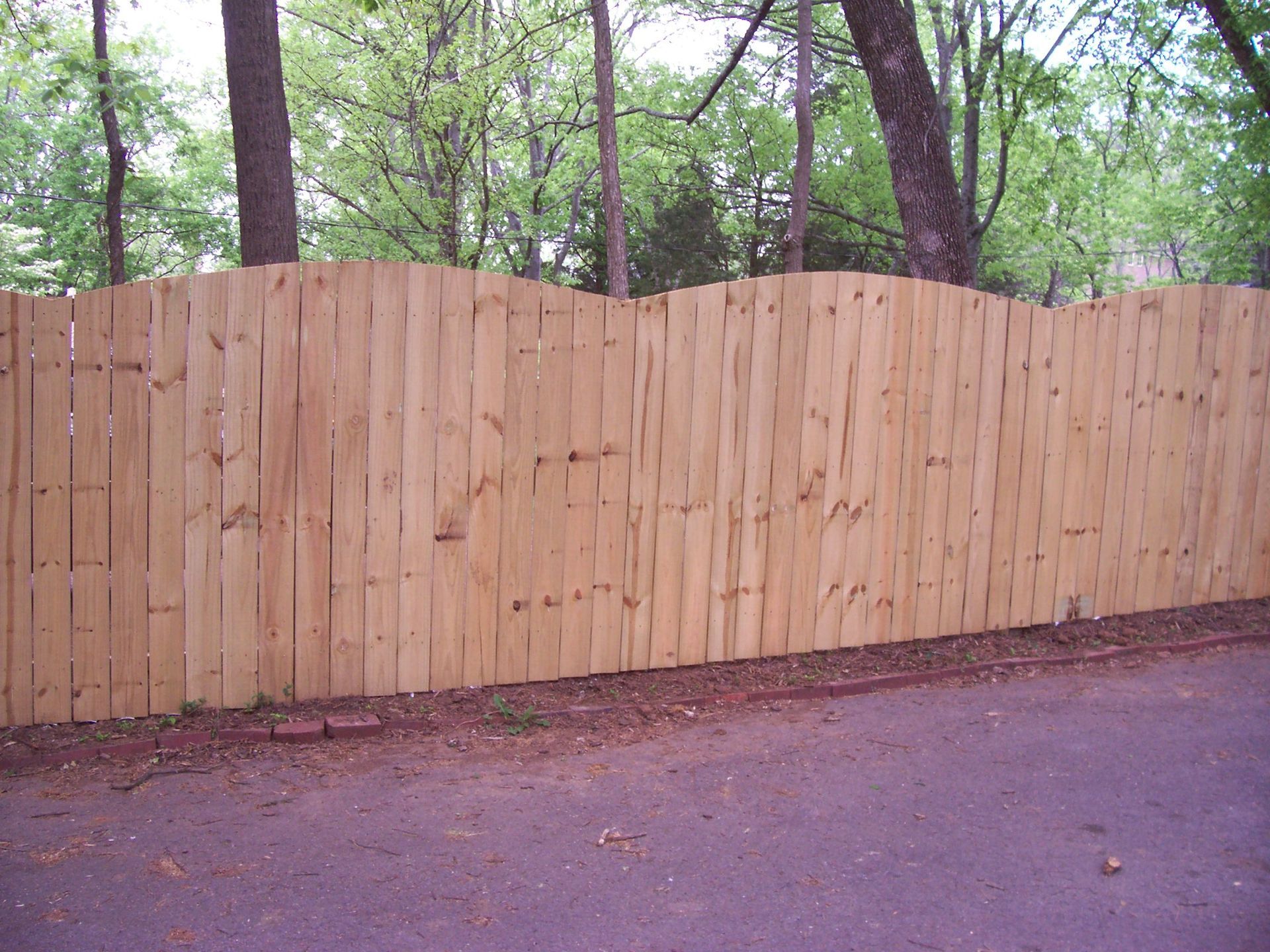 A light wood, scalloped-top privacy fence stands along a paved path in front of trees.