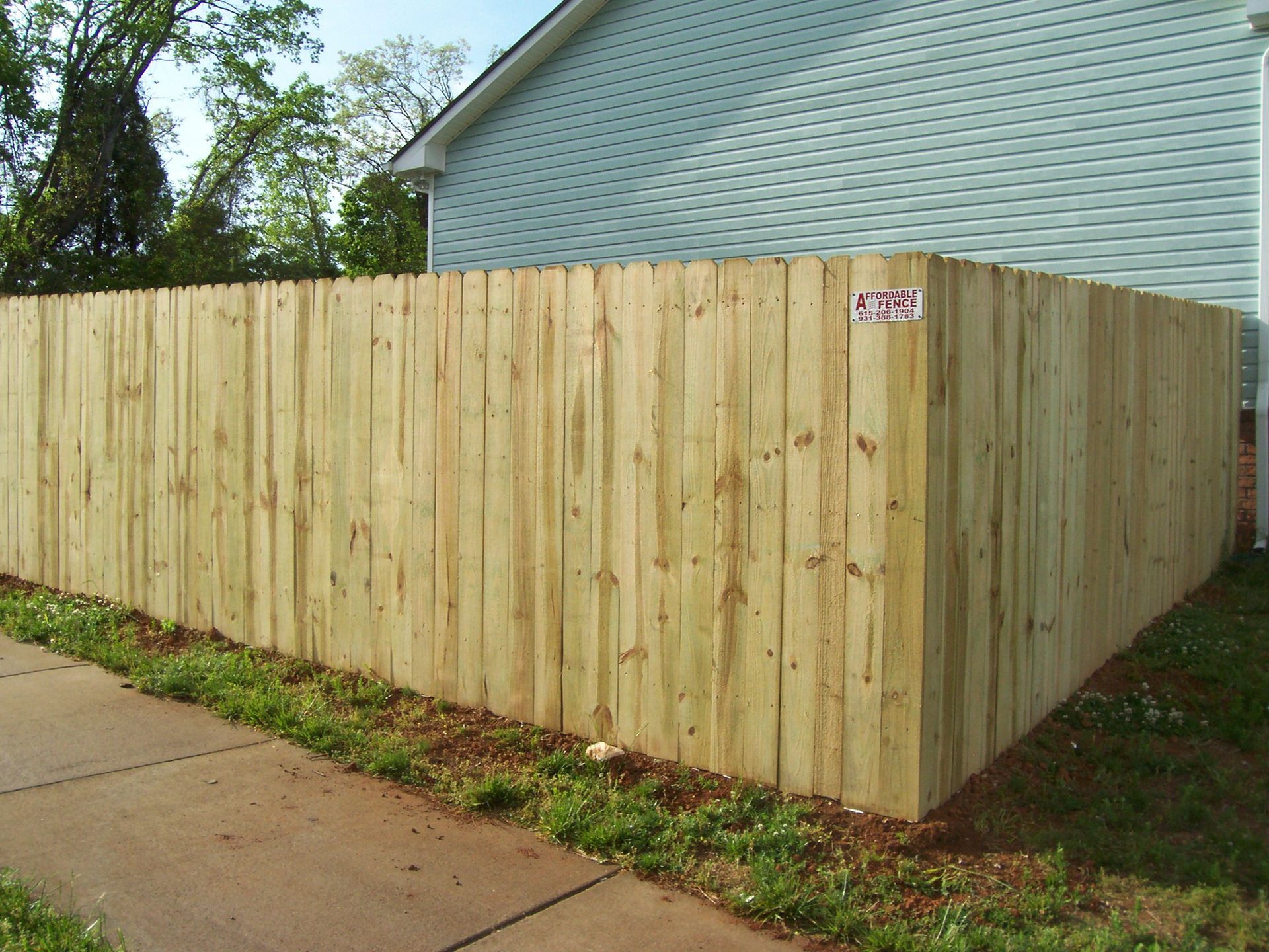 A new, light-colored wooden privacy fence stands in a grassy yard beside a light blue house near a sidewalk.