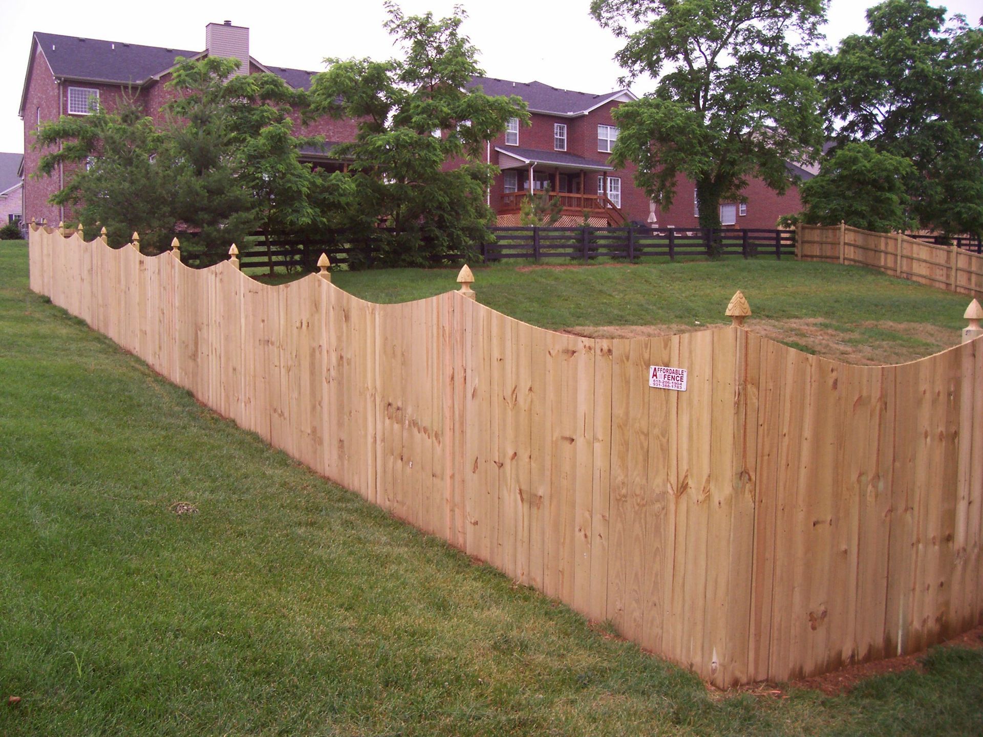 A new wooden scalloped-top privacy fence installed in a suburban backyard with brick houses in the background.