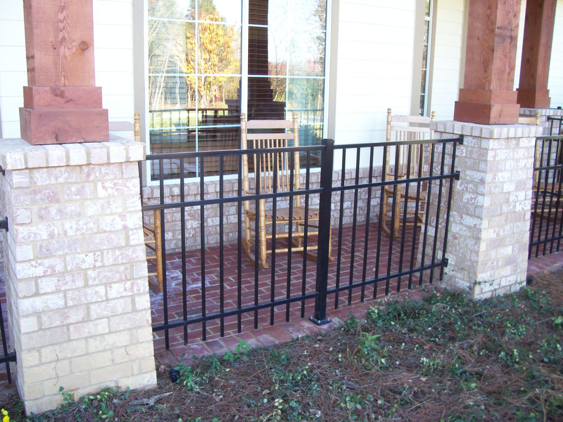 Black metal railing on a brick porch between stone columns with two rocking chairs visible through a large window.
