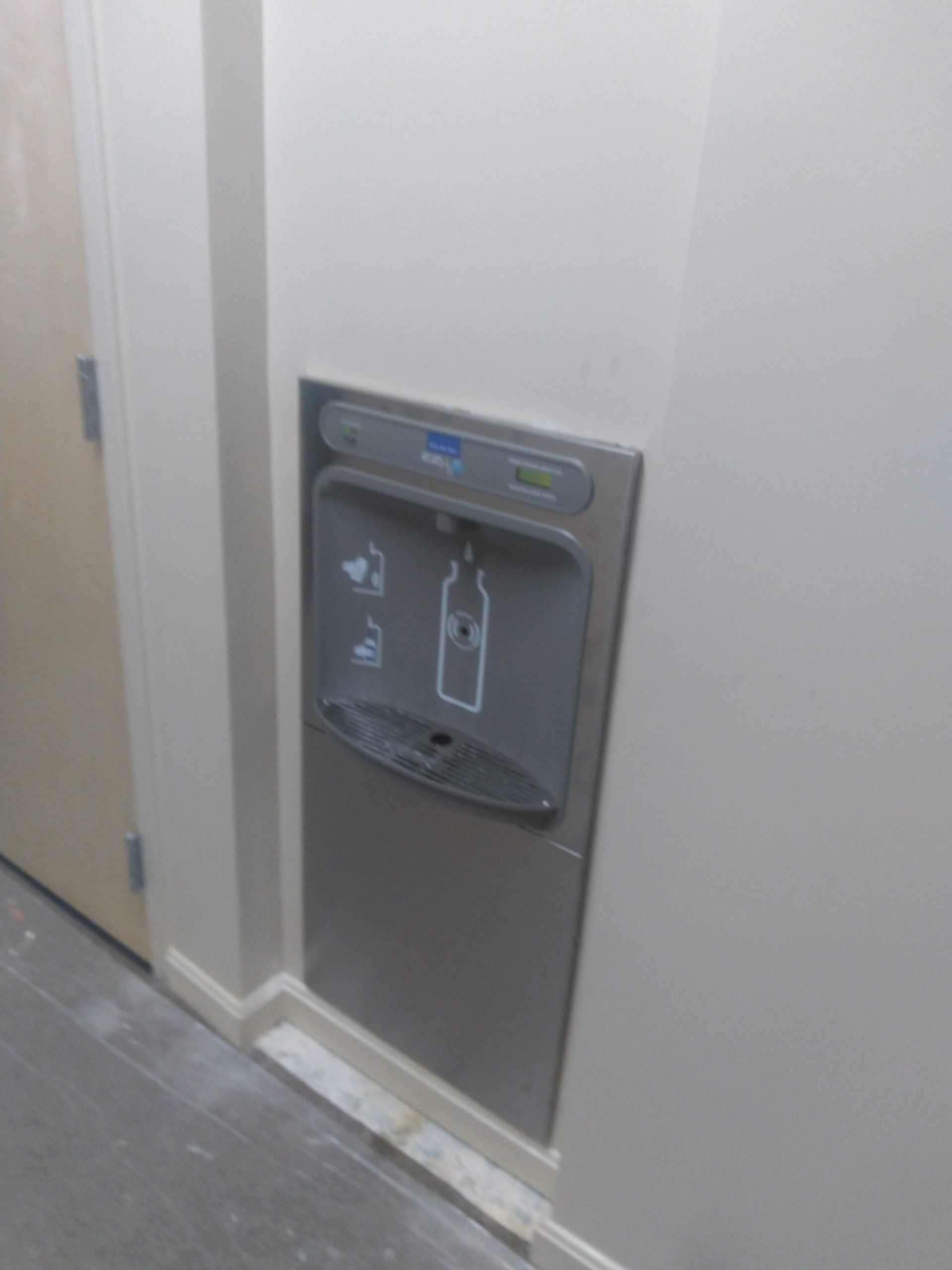 Stainless steel water bottle filling station on a beige wall. Near a door on the left.