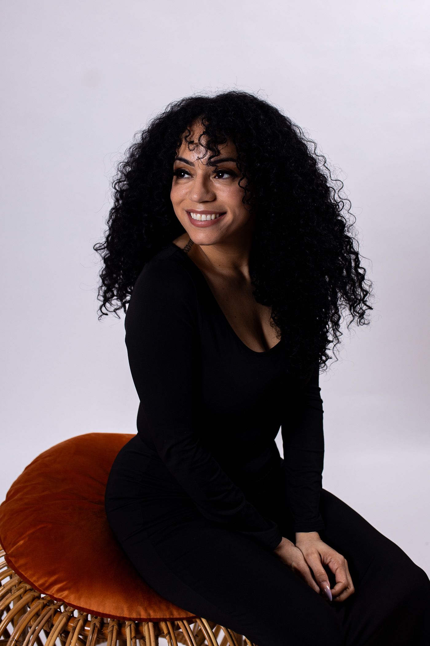 Smiling person in a black outfit seated on an orange chair against a light gray background - headshot session