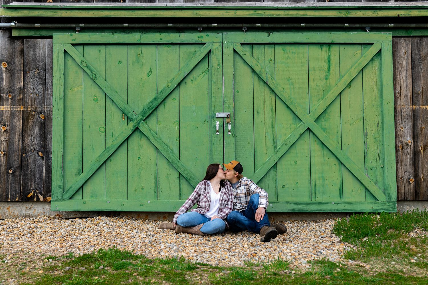 Couples sitting in front of green doors for barn, kissing engagement session