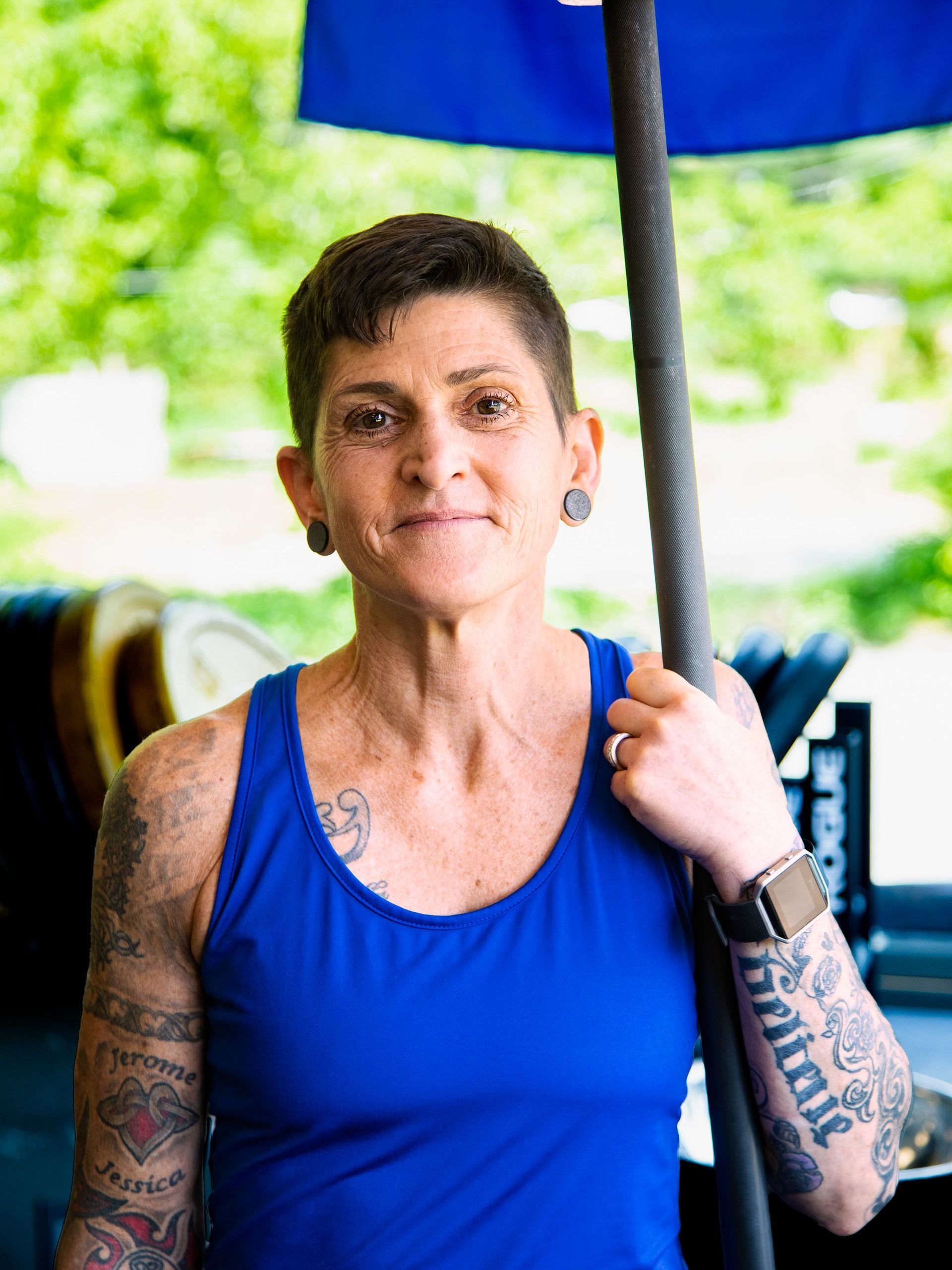 Photography MA -A woman in a blue tank top with tattoos on her arms is holding a barbell.