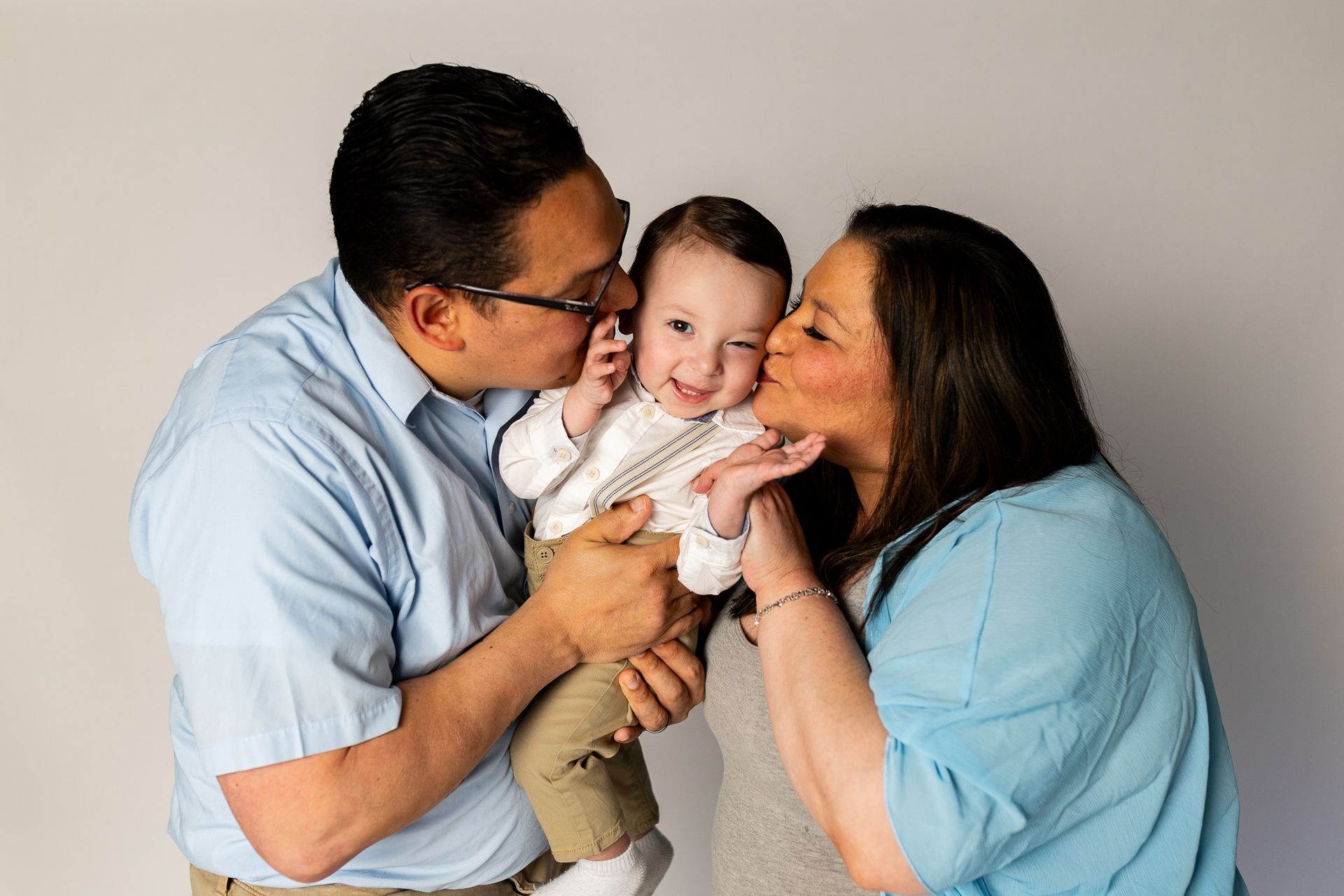 Three people kissing a smiling baby against a plain background - family photos