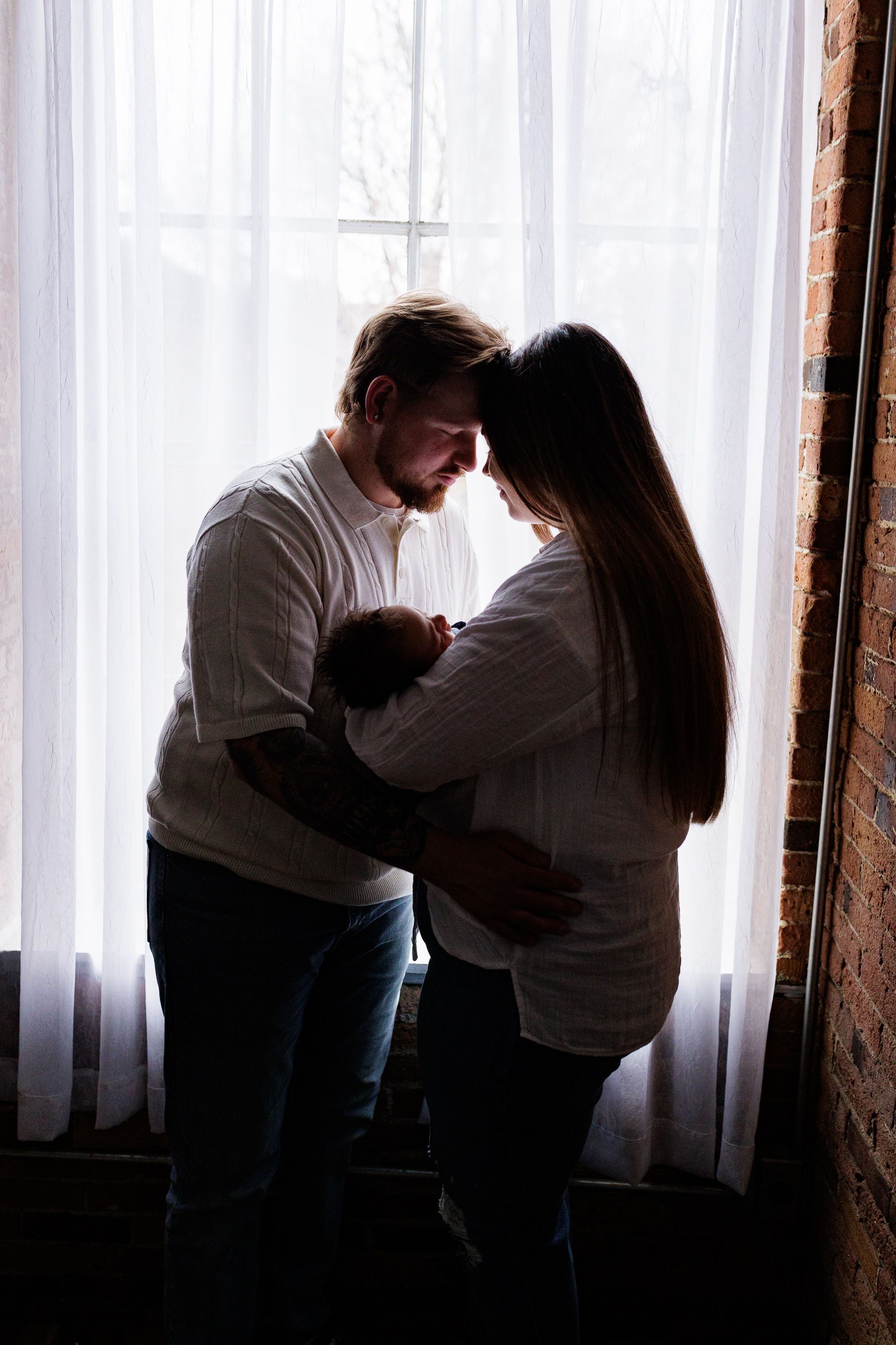 Newborn photography parents snuggling by window while holding baby