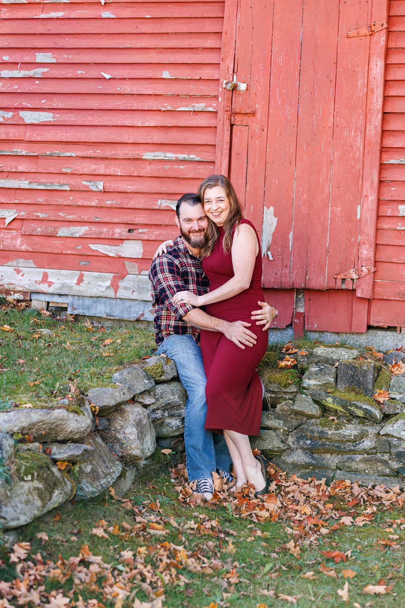 Couple embracing in front of a weathered red barn with stone foundation and fallen autumn leaves - family photos