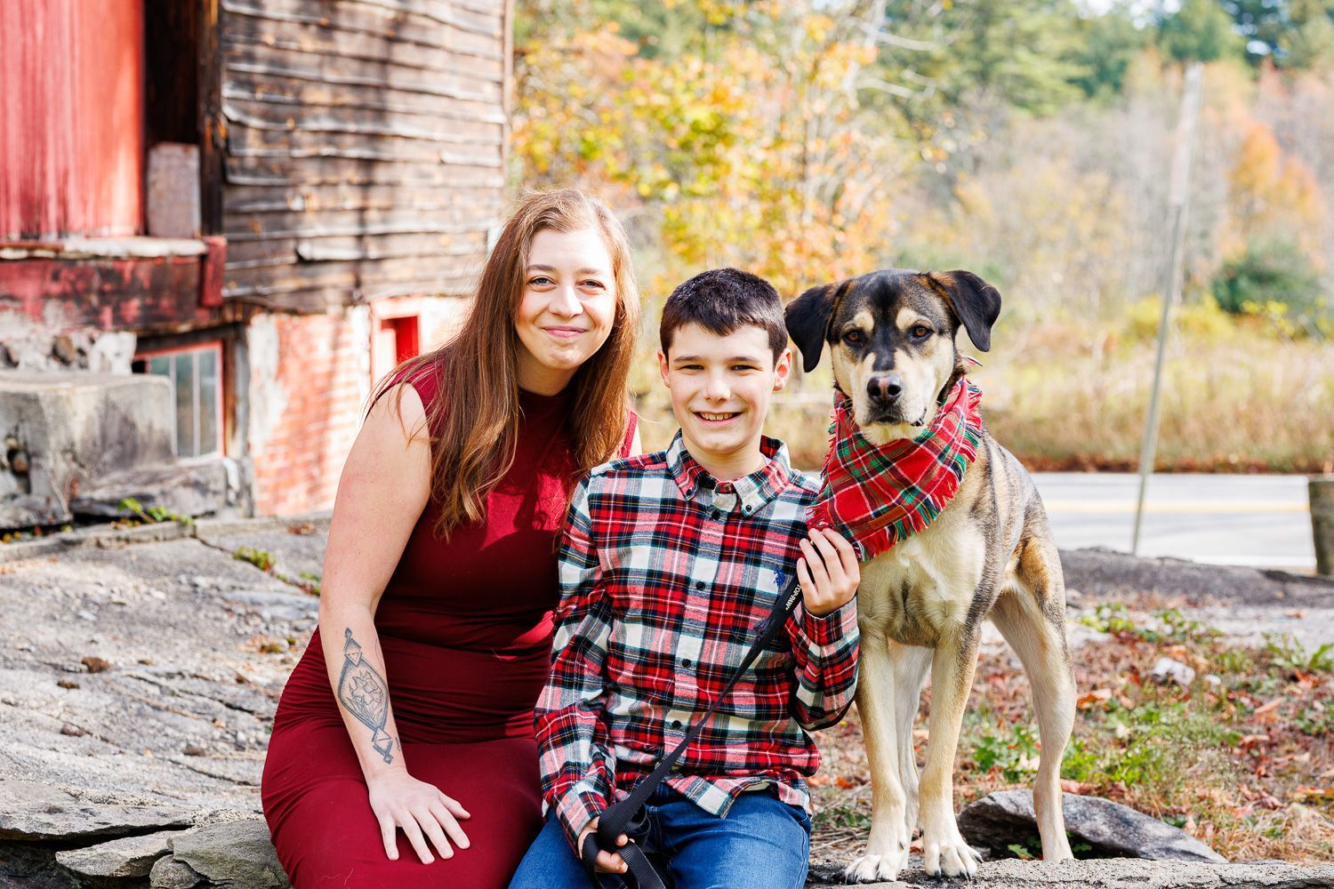 Smiling woman and boy in red plaid sit beside a dog wearing a red bandana outdoors near a barn - family photos