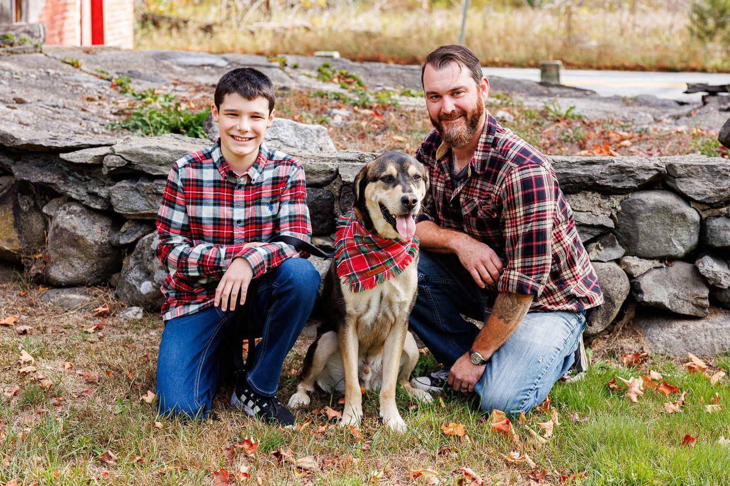 Boy and bearded man kneel beside a dog in red bandanas outdoors by a stone wall - family photos