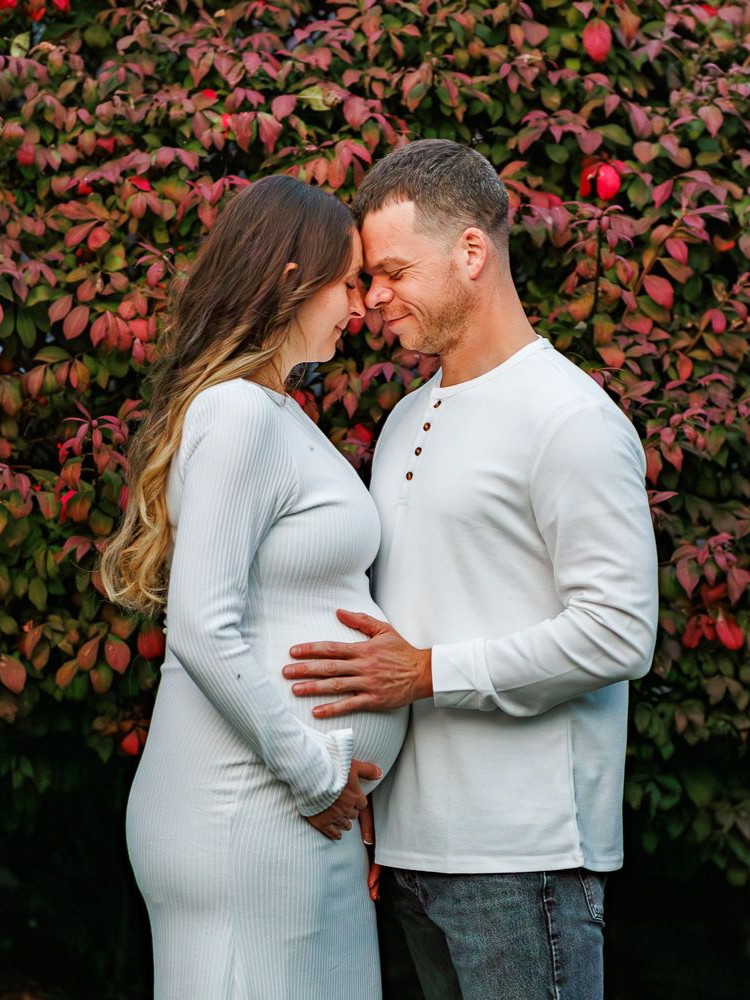 A man and a pregnant woman are standing in front of a tree with white flowers.