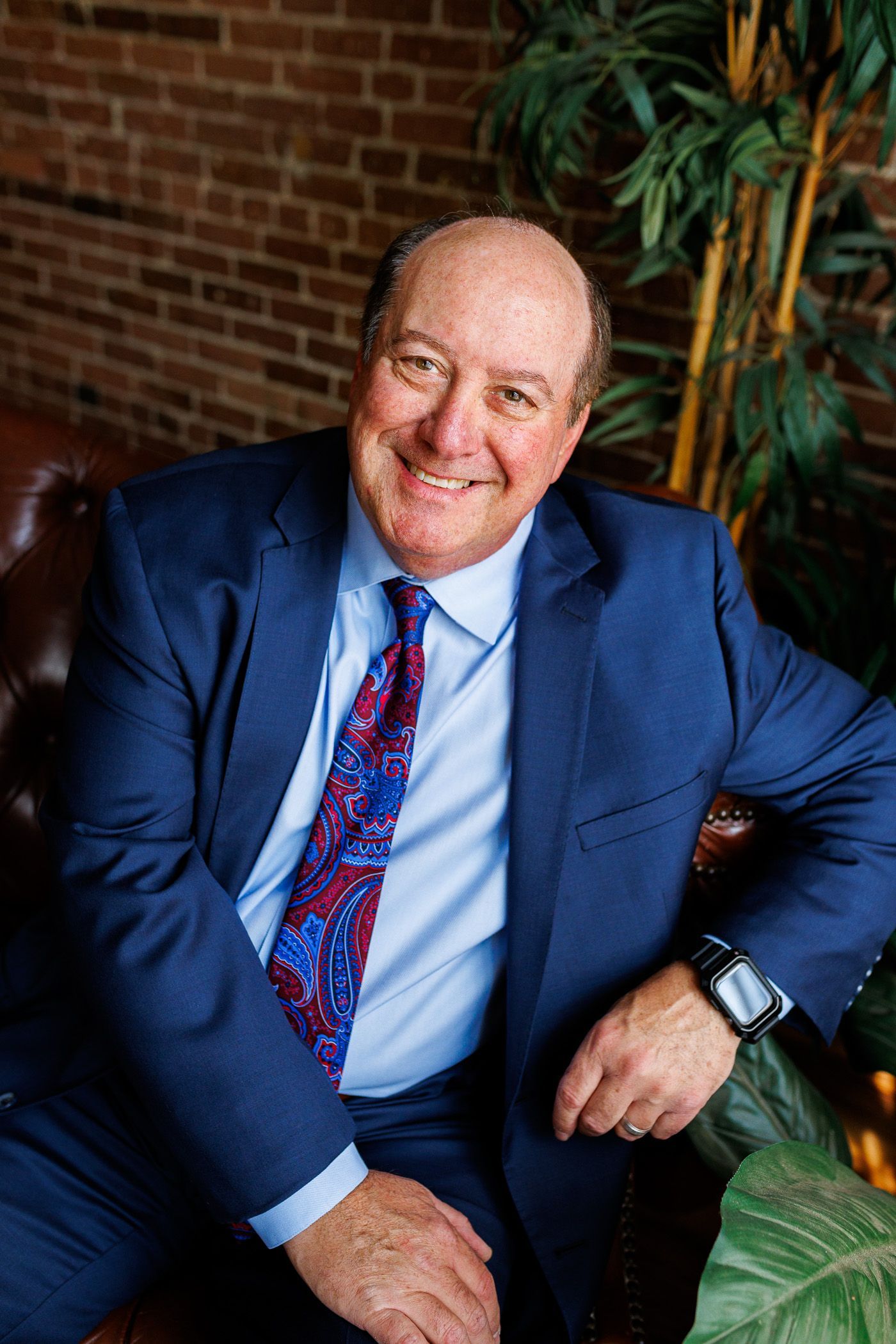 Smiling man in a blue suit seated indoors beside a brick wall and plants - headshot session