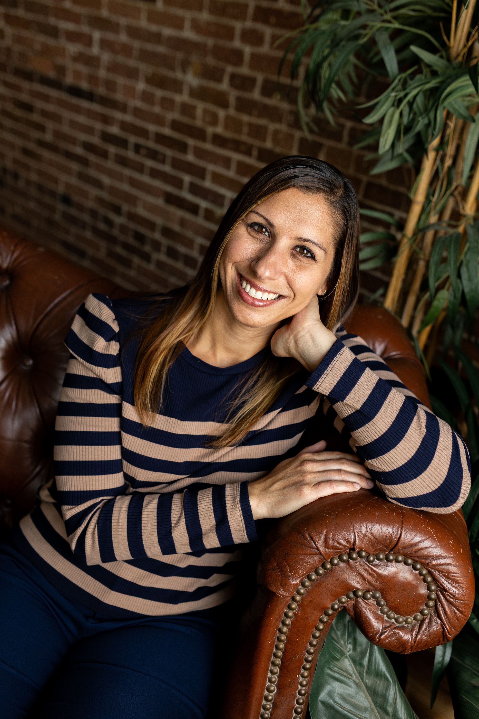 Smiling woman in a striped sweater sitting on a brown leather sofa against a brick wall - headshot session
