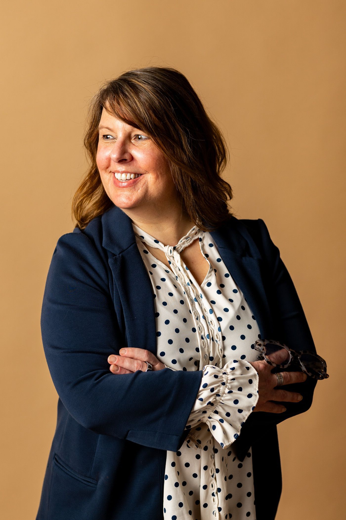 Smiling person in a navy blazer and patterned blouse, standing with arms crossed against a tan background - headshot session