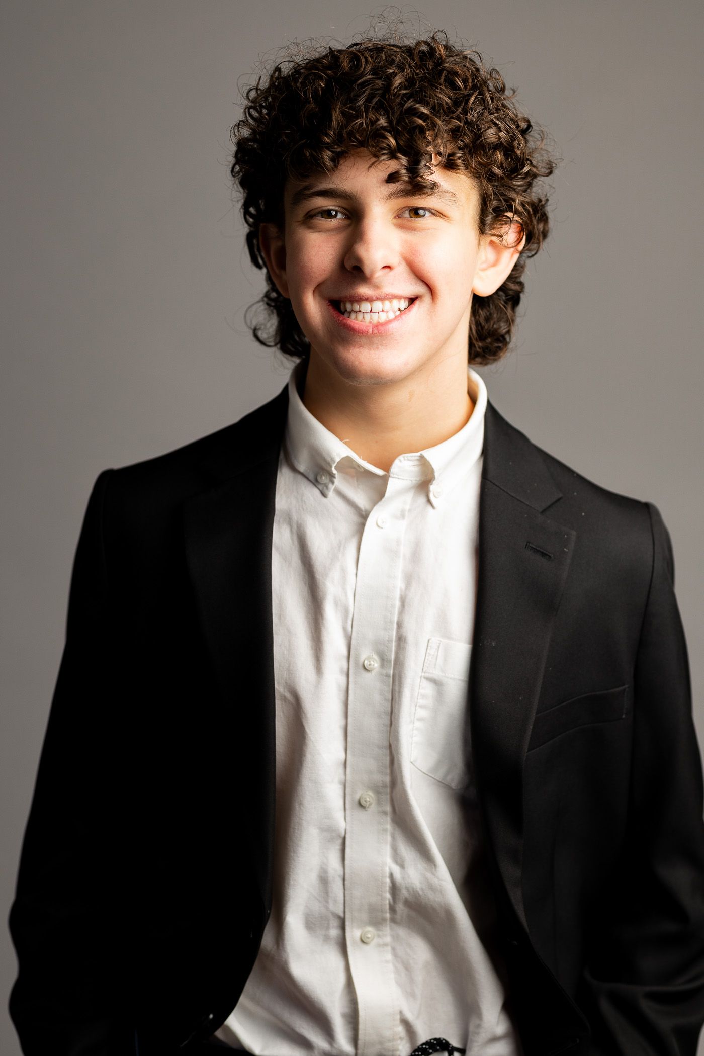 Smiling person with curly hair in a black blazer and white shirt against a plain gray background - headshot session