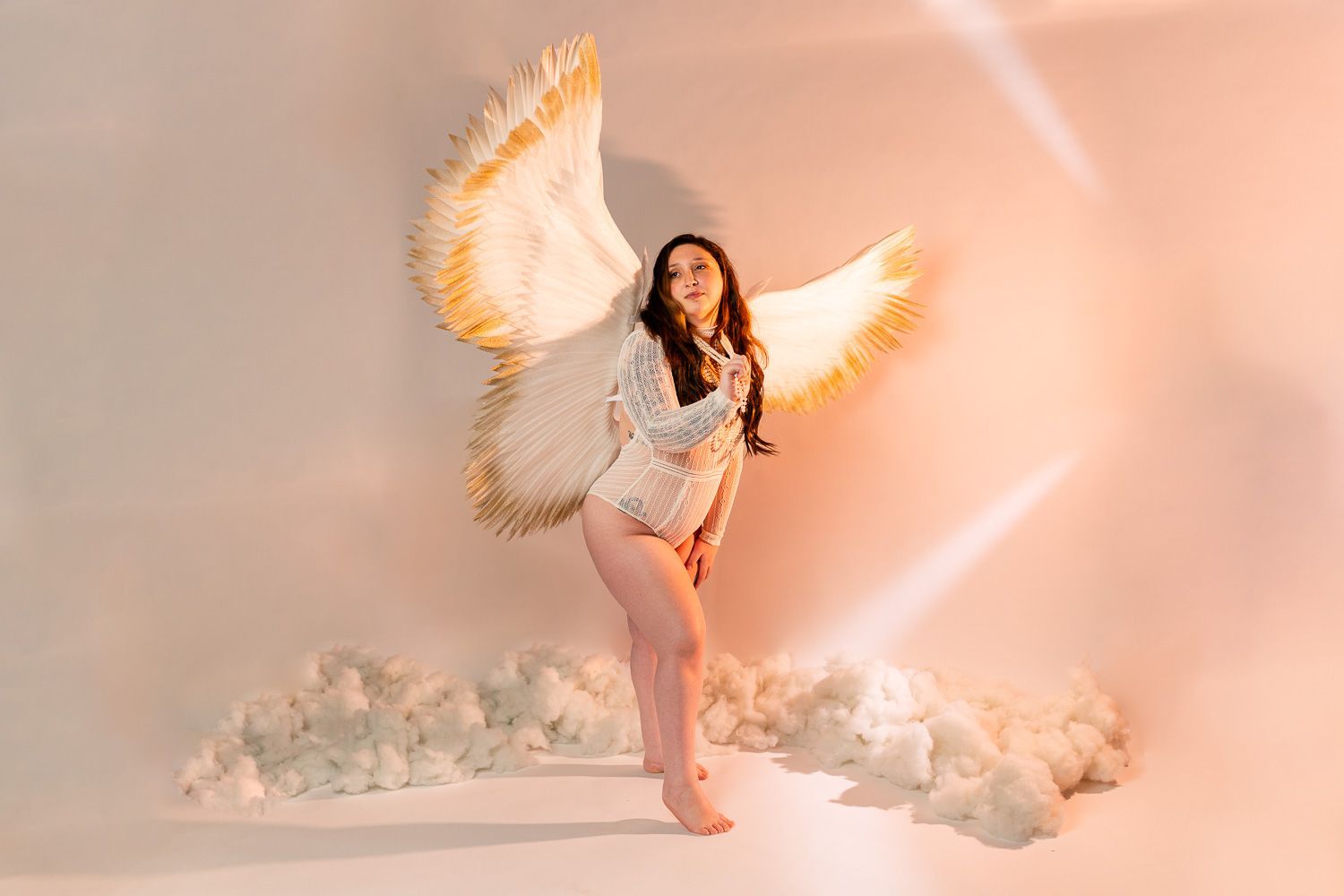 Woman posing in lingerie for boudoir session posing with angel wings on a peach background