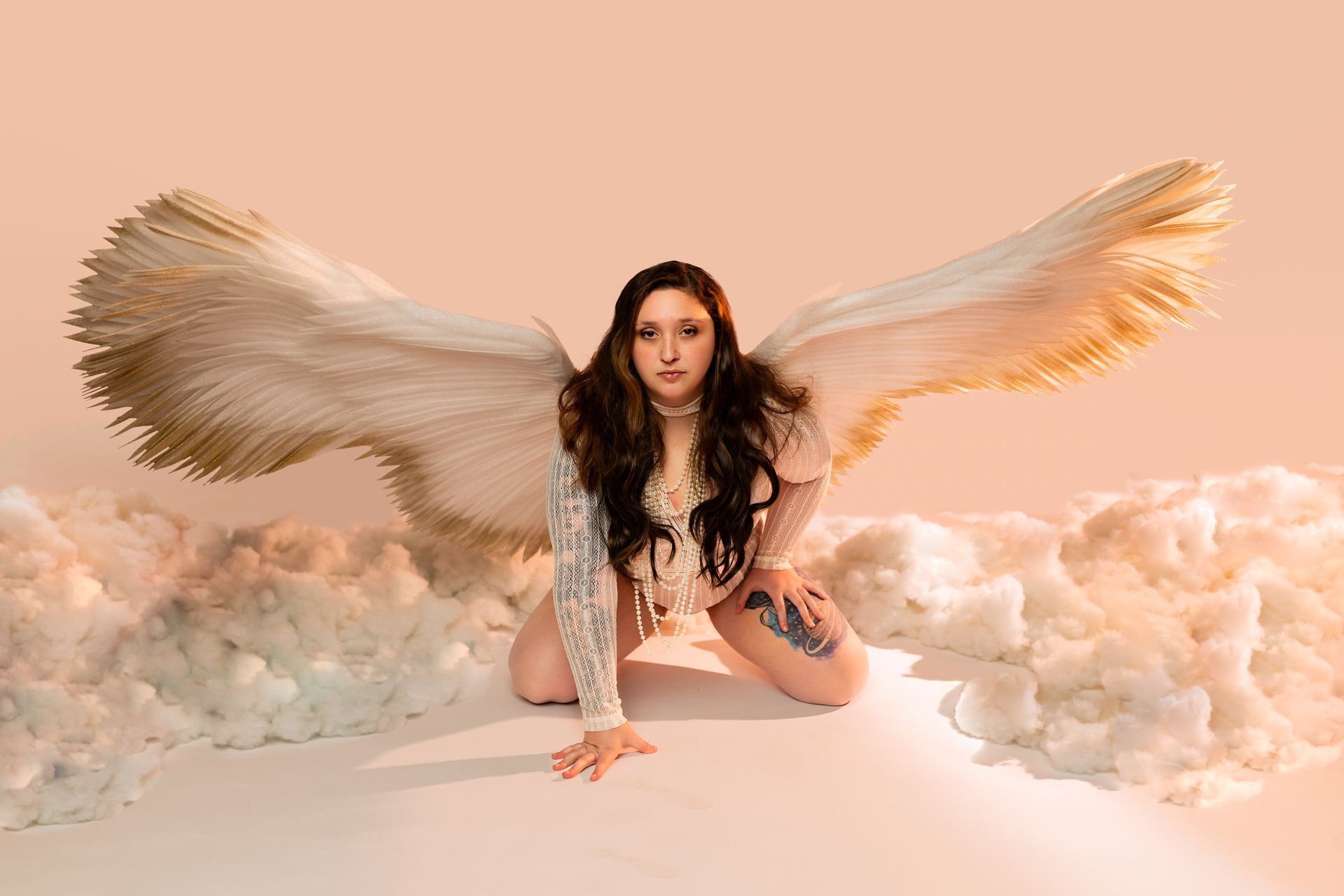 Woman posing in lingerie for boudoir session with angel wings on a peach background with clouds 
