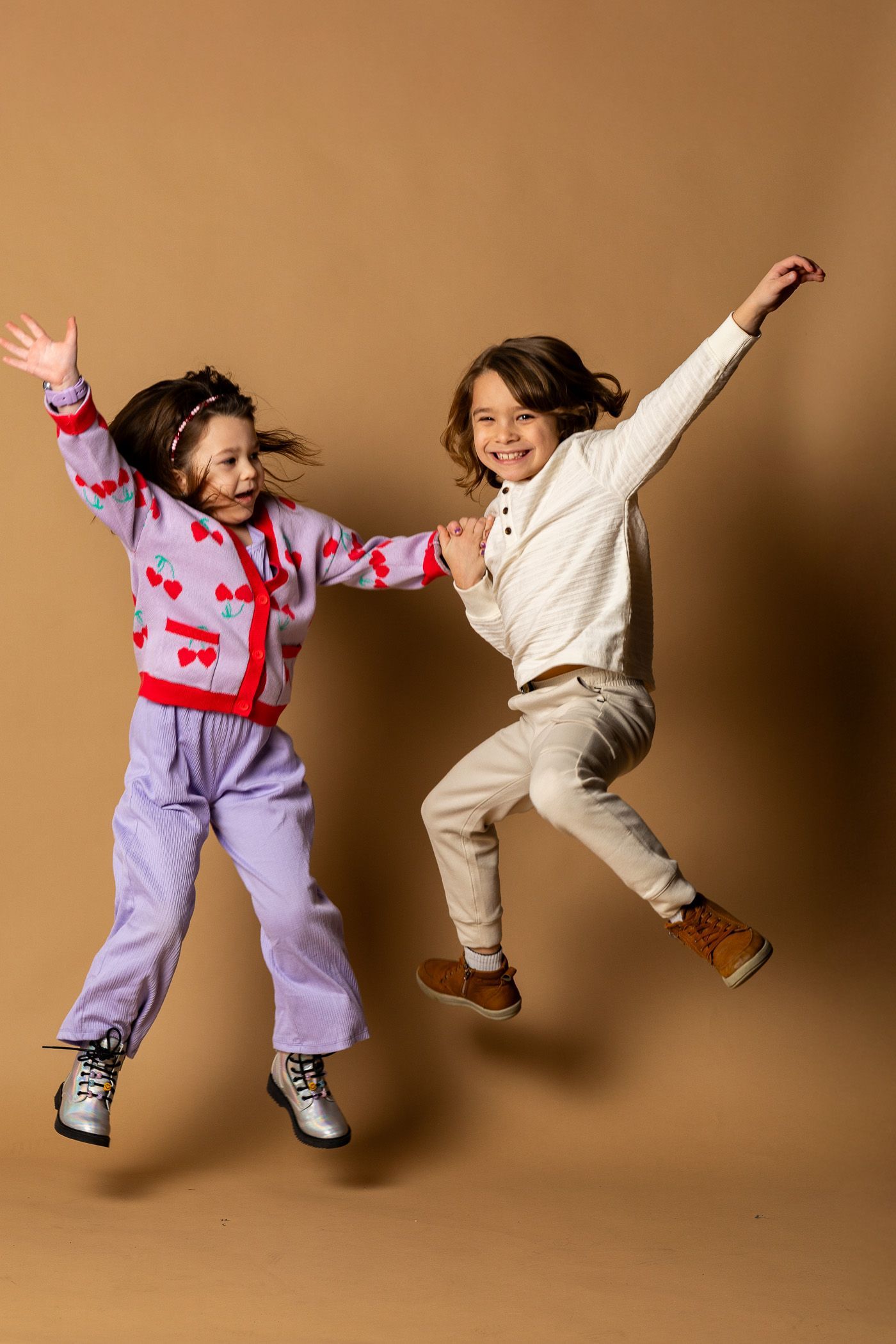 Two children jumping with arms raised against a tan studio backdrop - family photos