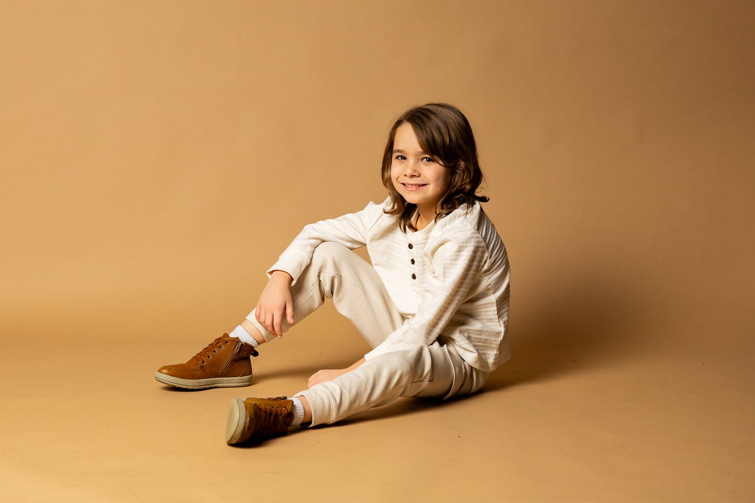 Young child sitting on a tan backdrop, smiling in a white outfit with brown boots. - family photos