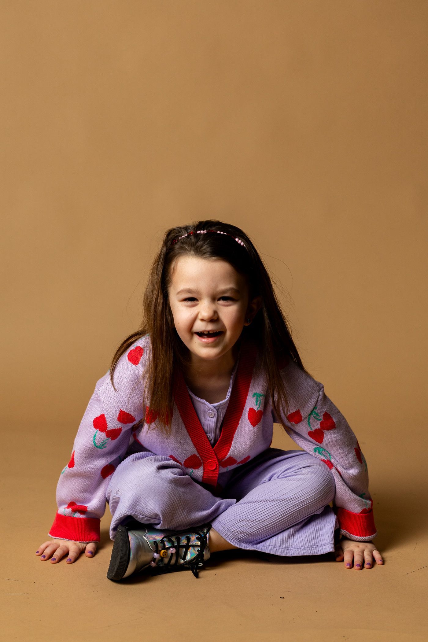 Young child sitting cross-legged on a studio floor, smiling in a red heart sweater and purple pants. - family photos
