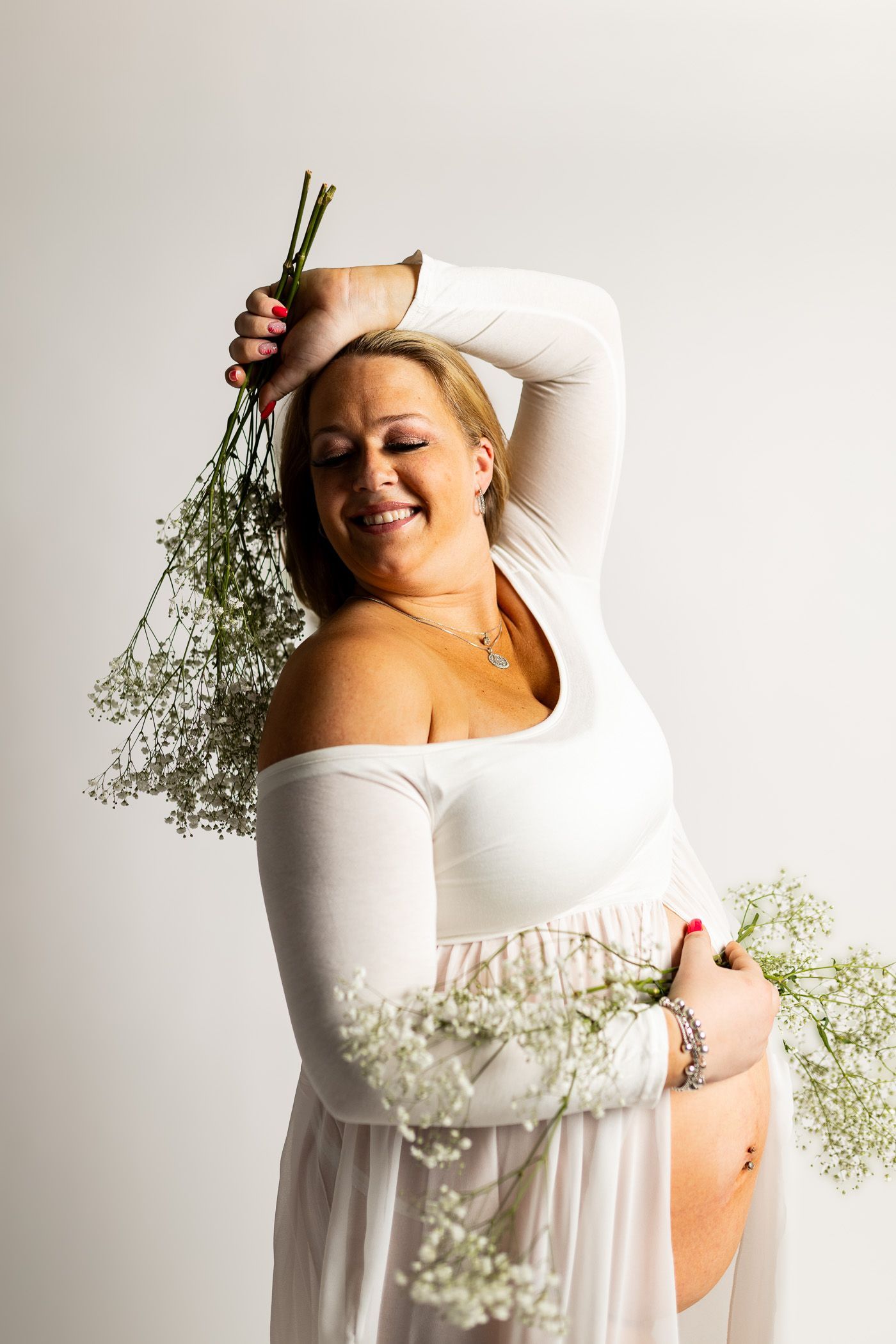 Maternity photography mom posing in white dress on white studio background with flowers